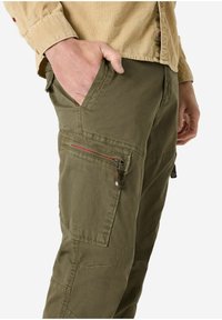 Olive green cargo pants made from sturdy fabric. Features side pockets, one with a red zipper, and visible stitching details.