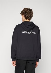 Černá mikina s kapucí, na zádech velký bílý nápis "INTERNATIONAL ENGLAND." Žebrované manžety a lem.