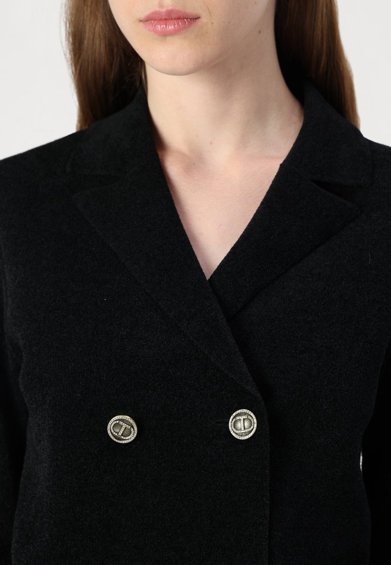 Schwarzes, doppelt knöpfbares Blazer aus strukturiertem Wolle mit scharfen Revers und metallischen Knopfakzenten. Nahaufnahme, die den Stoff und die Verschlüsse hervorhebt.