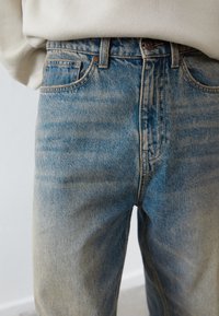 Primer plano de unos vaqueros de mezclilla azul desgastados, llevados con una blusa de color claro, que muestra los bolsillos delanteros y el cierre de botón.