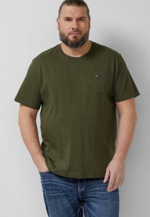 Mężczyzna z brodą, ubrany w oliwkowozielony t-shirt z krótkim rękawem i kieszenią na piersi oraz niebieskie dżinsy, stojący na jednolitym szarym tle.