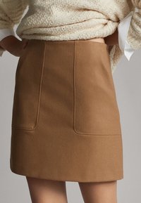 Jupe mini marron en tissu texturé avec deux poches horizontales et une coupe ajustée. Elle est associée à un haut en tricot de couleur crème.