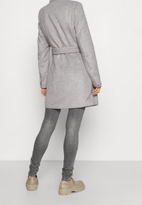 Persona che indossa un cappotto cinquetta grigio chiaro con cintura, jeans skinny grigio scuro e mocassini chunky beige, in piedi rivolto verso un muro bianco.