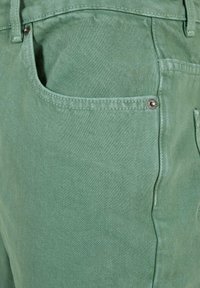 Žalios džinsinės kelnės, turinčios minkštą tekstūrą, su šonine kišene, metalinėmis klinkėmis ir tvarkingomis siūlėmis.