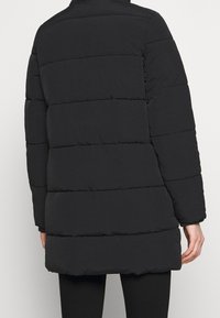 Osoba nosící černou péřovou bundu do poloviny stehna, zobrazená zezadu, stojící proti jednobarevnému světlému pozadí.