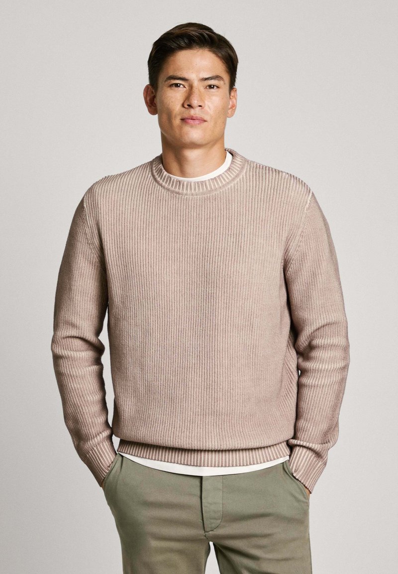 Vaalean beige ribbetty neule pyöreällä kaula-aukolla, jossa on tekstuuri ja pitkät hihat, valkoisen paidan päällä, yhdistetty vihreisiin housuihin.