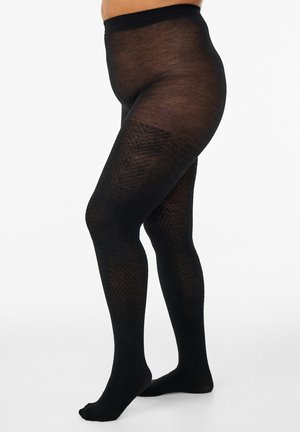 Person trägt schwarze, gemusterte blickdichte Strumpfhose, steht mit überkreuzten Beinen, gezeigt vom Rumpf bis zu den Füßen vor schlichtem Hintergrund.