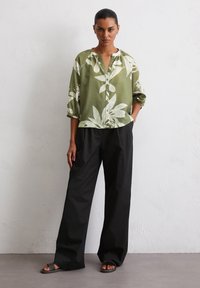 Green linen shirt with white leaf print, button-down design, and roll-up sleeves. Paired with black wide-leg trousers and flat sandals.