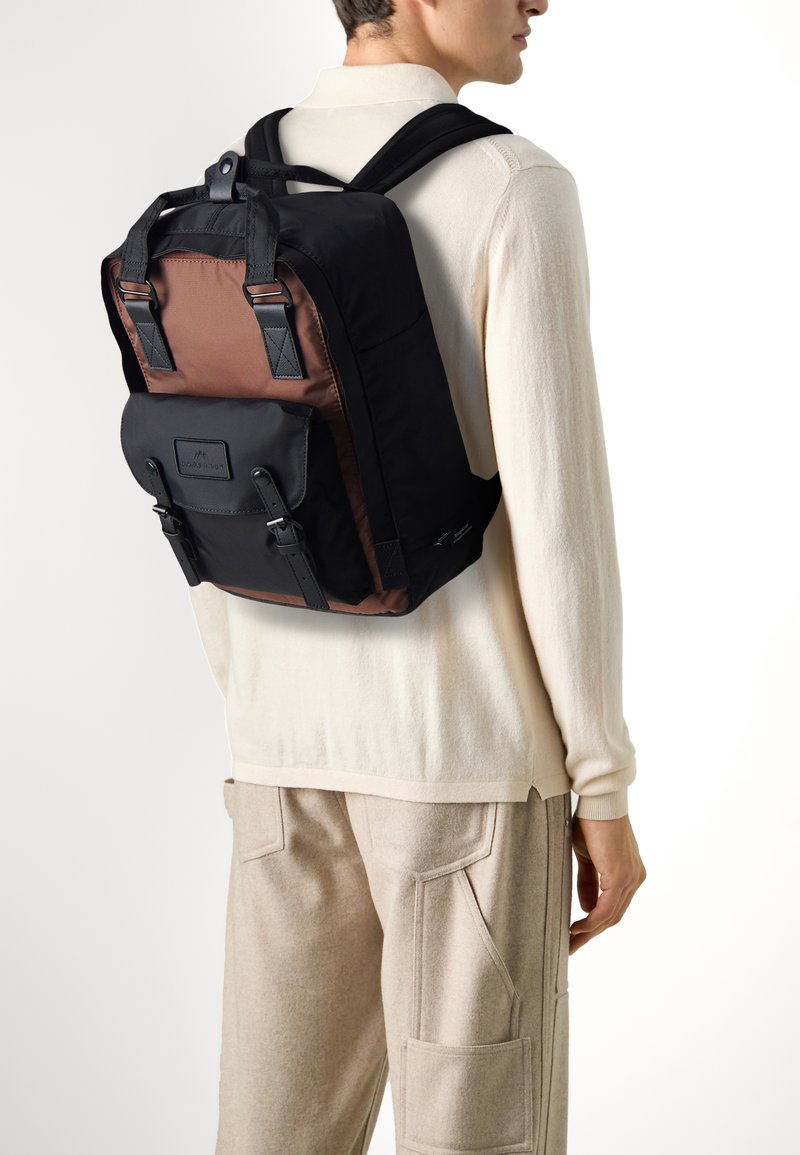 Schwarzer und brauner Stoff-Rucksack mit einer Vorderklappe, verstellbaren Riemen und einem strukturierten Design. Verfügt über Schnallen und kontrastierende Materialien.