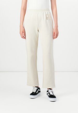 Personne portant un pantalon de survêtement couleur crème avec le logo "GAP" sur la taille gauche, associé à des baskets noires et blanches, se tenant sur un sol blanc.