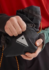 Schwarze, packbare Jacke mit grauem Logo, in einer Hand gehalten. Sie hat eine strukturierte Oberfläche und eine leuchtend orange-gelbe Kordel zum Schließen.