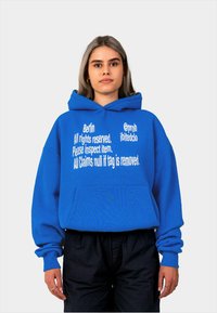 Felpa con cappuccio blu con testo bianco: "Berlino, Tutti i diritti riservati. Si prega di ispezionare l'articolo. Tutte le contestazioni sono nulle se l'etichetta viene rimossa." Tasca frontale.