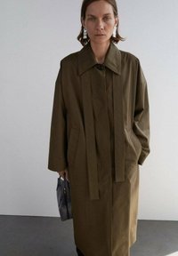 Cappotto lungo marrone con colletto, dotato di due tasche frontali e pannelli verticali. Il modello tiene in mano una borsa nera, con orecchini d'argento visibili.
