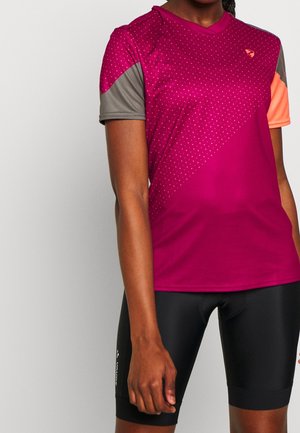 Kobieta ubrana w magentowy krótki sportowy T-shirt z szarymi i pomarańczowymi rękawami oraz czarne szorty rowerowe, stojąca na jednolitym tle.