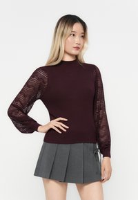 Bordo rebrasti pleteni džemper s rukavima na napuhavanje s čipkastim uzorkom, uparen sivi plisirani suknja s detaljem vezanja sa strane.