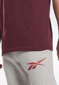 Camiseta marrón y pantalones de chándal grises. Los pantalones tienen un logotipo rojo en el muslo izquierdo. El tejido parece suave y tiene un corte relajado.