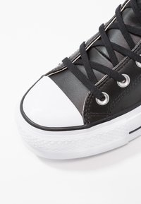 Czarne skórzane sneakersy z białą gumową osłoną na palce, czarnymi sznurowadłami, srebrnymi oczkami i teksturowaną białą podeszwą. Po bokach biegną przeszycia, które dodają szczegółów.