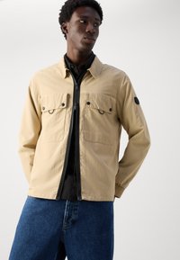 Beige jack met rits aan de voorkant, twee borstzakken met kleppen, elastische manchetten en een ronde patch op de linkermouw, gedragen over een zwart shirt.