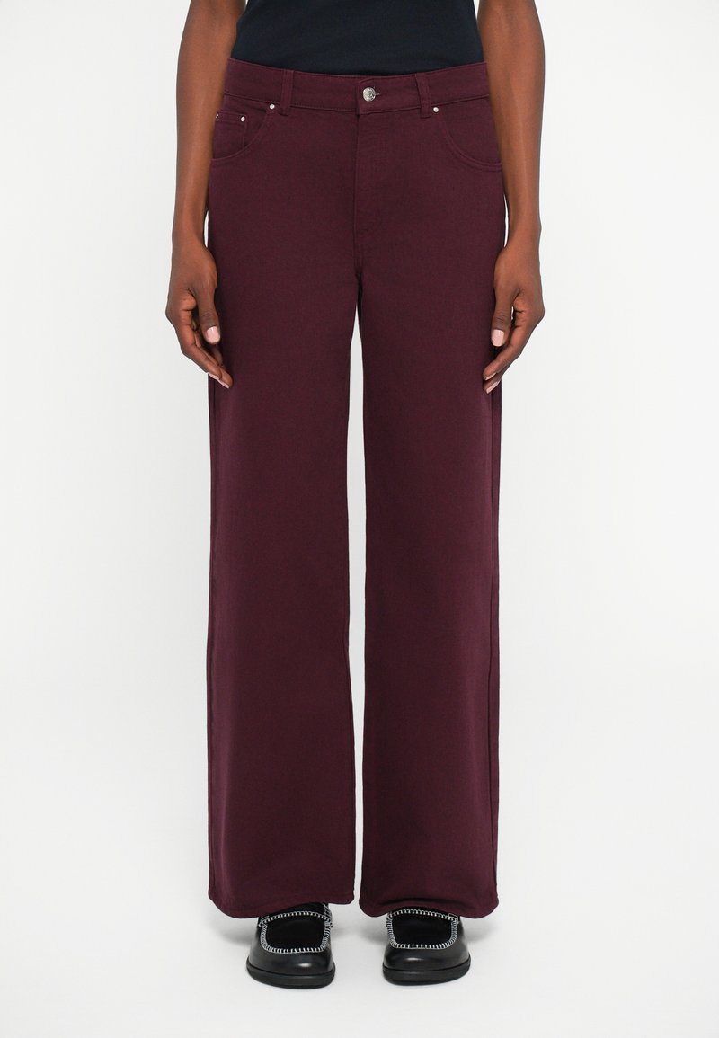 Pantalones de pierna ancha burdeos en una tela suave con una cremallera de botones, dos bolsillos frontales y un ajuste relajado, combinados con zapatos negros.