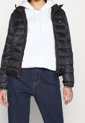 Veste matelassée noire avec zip, dotée d'une capuche et d'accents rouges. Portée sur un sweat à capuche blanc et un pantalon en denim foncé. Texture lisse.