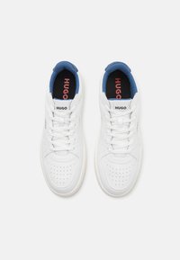 White sneakers with a smooth leather upper, perforated toe, blue accents on the collar, and black insoles featuring the "HUGO" logo in red.