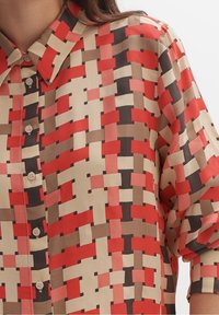 Camicia multicolore con un motivo geometrico in rosso, beige e marrone. Maniche corte e chiusura frontale con bottoni e colletto classico.