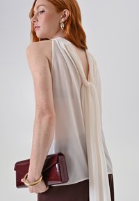 Bianchetta senza maniche con dettaglio drappeggiato sul retro, abbinata a una pochette bordeaux tenuta sotto un bracciale dorato. Tessuto morbido, design minimalista.