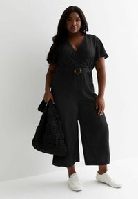 Svart, omslagstyp jumpsuit med korta ärmar, bälte i midjan med en rund spänne, tillsammans med en texturerad svart tote och vita sneakers.