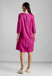 Frau mit dunklen Haaren im Pferdeschwanz, die ein lockeres, knielanges magentafarbenes Kleid mit 3/4-Ärmeln und weiße Turnschuhe trägt, steht vor einem einfarbigen Hintergrund.