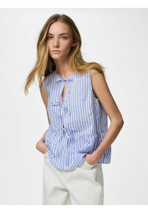 Jeune femme portant un chemisier sans manches rayé bleu et blanc avec des nœuds noués, pantalon blanc, mains dans les poches, debout devant un fond uni.