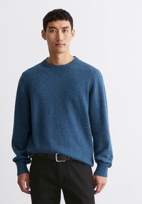 Blauer Strickpullover mit strukturiertem Aussehen, rundem Halsausschnitt und gerippten Bündchen, getragen über schwarzen Hosen.