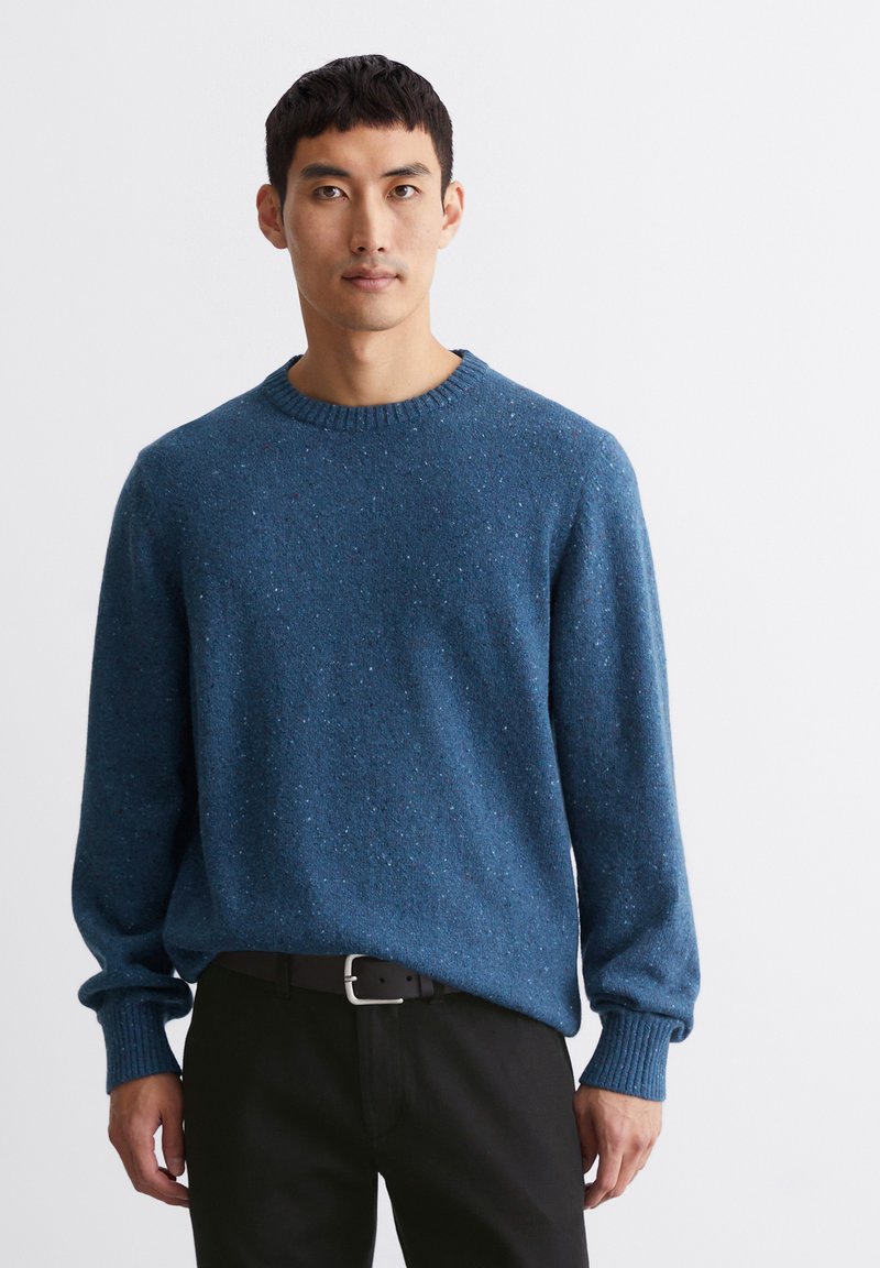 Blauer Strickpullover mit strukturiertem Aussehen, rundem Halsausschnitt und gerippten Bündchen, getragen über schwarzen Hosen.