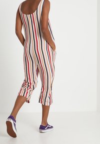 Combinaison rayée avec un dos plongeant, présentant des lignes verticales en rouge, bleu et beige. Longueur courte et poches latérales. Associée à des baskets violettes.