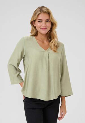Blouse légère, vert clair, avec un col en V et des manches trois-quarts. Présente un pli à l'avant et une coupe décontractée.