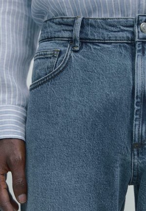 Zbliżenie osoby noszącej niebieskie dżinsy i jasnoniebieską koszulę w paski z długim rękawem, z ręką spoczywającą obok dżinsów.