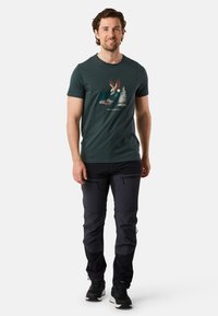 Mörkgrön bomull t-shirt med en grafik av en örn och träd, tillsammans med svarta byxor och svarta skor.