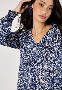 Blaue und cremefarbene gemusterte Bluse mit V-Ausschnitt, langen elastischen Ärmeln und Knopfdetails. Verfügt über ein welliges Paisleymuster und eine weiche Textur.