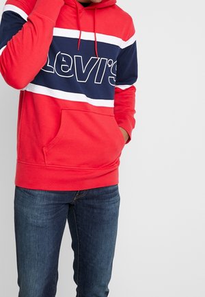 Mann trägt einen roten Levi’s Hoodie mit marineblauem und weißem Streifen, Hand in der Tasche und dunkelblaue Jeans vor schlichtem Hintergrund.