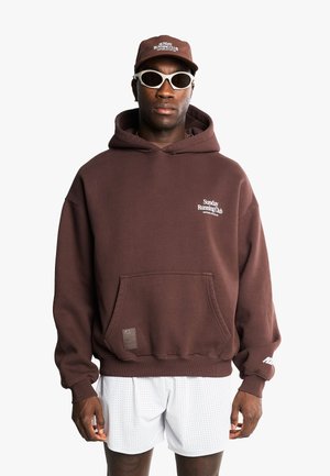Homme portant un sweat à capuche et une casquette marrons Sunday Running Club, des lunettes de soleil blanches futuristes et un short blanc perforé, debout sur fond blanc.