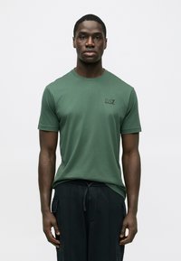 Zöld rövid ujjú pamut póló, kerek nyakkivágással és egy kis fekete logóval a mellkason. Feket nadrággal viselve.