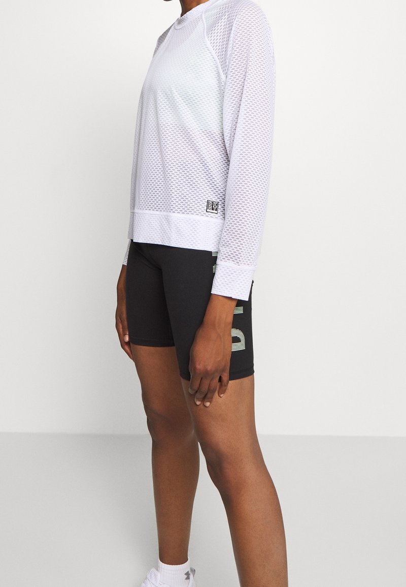 Weißes Mesh-Sportoberteil mit langen Ärmeln und entspanntem Schnitt, kombiniert mit schwarzen Shorts mit grünen grafischen Akzenten. Texturiertes und atmungsaktives Material.