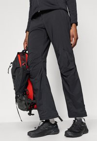 Pantalones exteriores negros hechos de material ligero, con un corte ajustado y bolsillos de carga, combinados con zapatos de senderismo negros y una mochila con ribetes rojos.