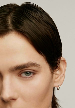 Pendiente geométrico plateado con forma de V abierta. El acabado metálico suave y el diseño elegante complementan el pelo corto y oscuro de la modelo.