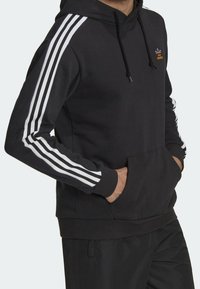 Svart hoodie med vita tre-randsdetaljer på ärmarna, känguruficka, dragsko i huven och broderad logotyp på bröstet.