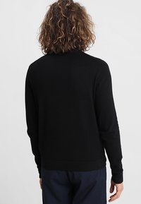 Personne avec des cheveux bouclés moyens portant un t-shirt noir à manches longues et un pantalon sombre, se tenant de dos contre un fond uni.