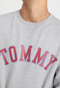 Sudadera gris con un escote redondeado y un gran texto rojo que dice "TOMMY JEANS" en una fuente estilizada en el pecho.