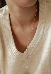 Długa naszyjnik w kolorze różowego złota z delikatnym łańcuszkiem i pojedynczym zawieszką w postaci perły, noszony na kremowym swetrze w prążki.