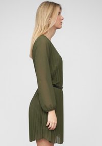 Zelené plisované šaty s dlouhými rukávy, páskem v pase a průhlednými detaily. Design zahrnuje jemnou, splývavou texturu.