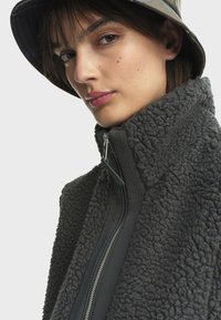 Veste en polaire gris foncé avec col montant, tissu texturé et fermeture éclair centrale. Le modèle porte un chapeau seau à motifs.