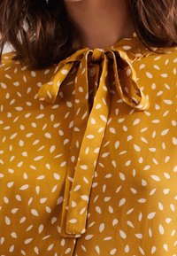 Mustard blouse with white leaf patterns, featuring a tied collar, button closure, and smooth fabric texture.